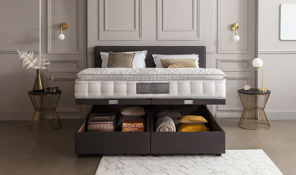 Lit avec matelas Savoy, tiroirs de rangement ouverts contenant des coussins et couvertures, tables de chevet modernes avec lampes dorées