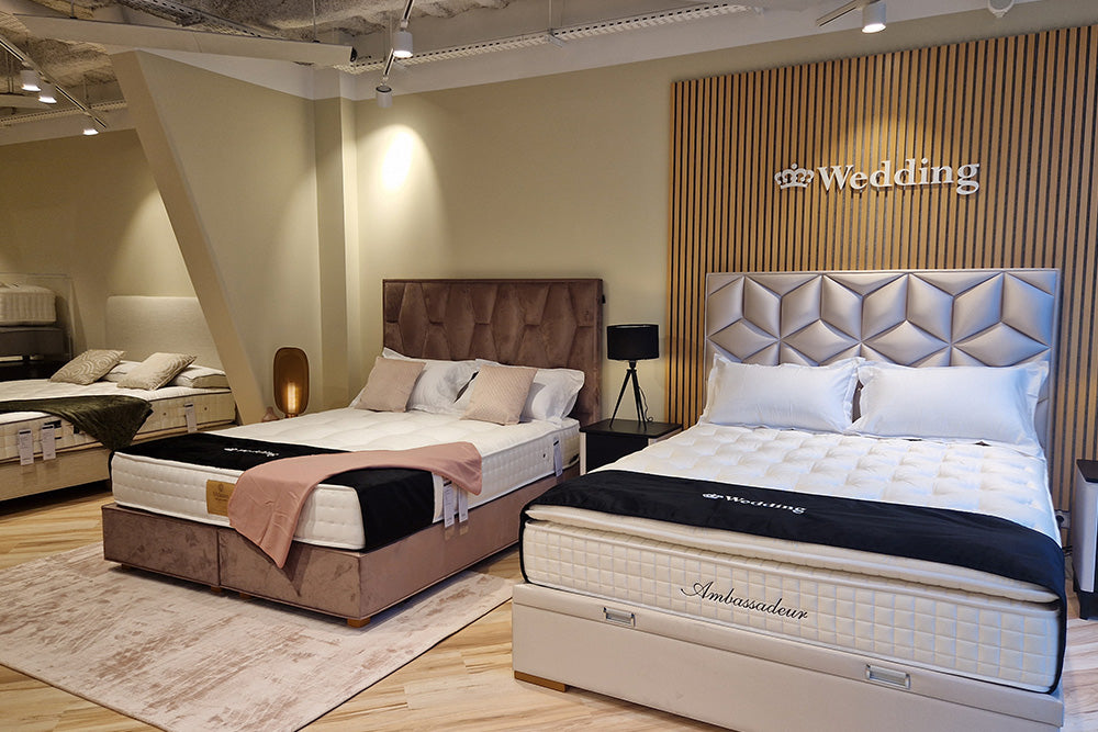 Deux lits luxueux exposés dans un showroom, avec des têtes de lit élégantes et des couvertures marquées "Wedding" et "Ambassadeur"