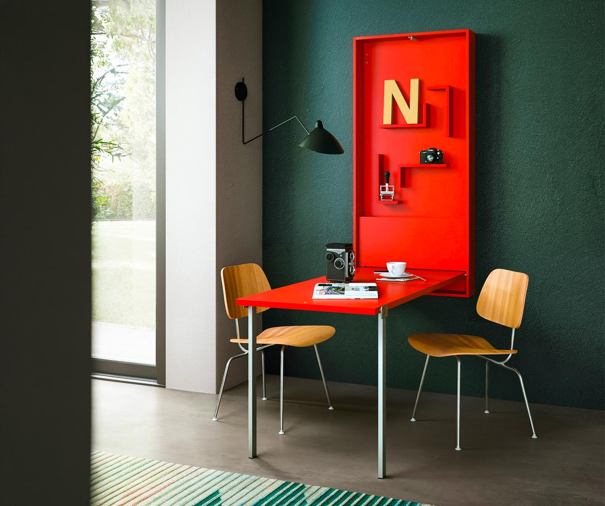Bureau WALLY +50 coloris & matériaux plié en table rouge fixée à un mur vert foncé, entourée de deux chaises en bois clair dans un intérieur lumineux avec lampe noire murale.