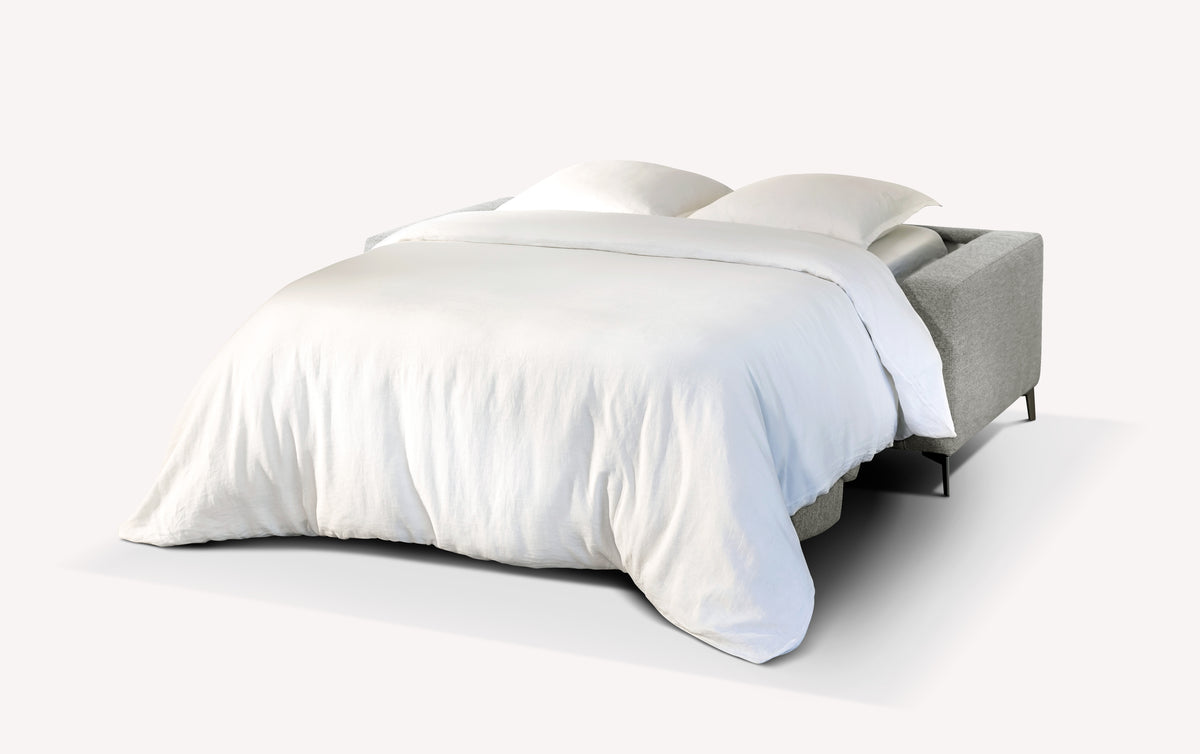 Lot canapé et pouf LISBONA en tissu gris clair transformé en canapé convertible avec un lit déplié et une literie blanche, offrant une solution élégante pour couchage quotidien.