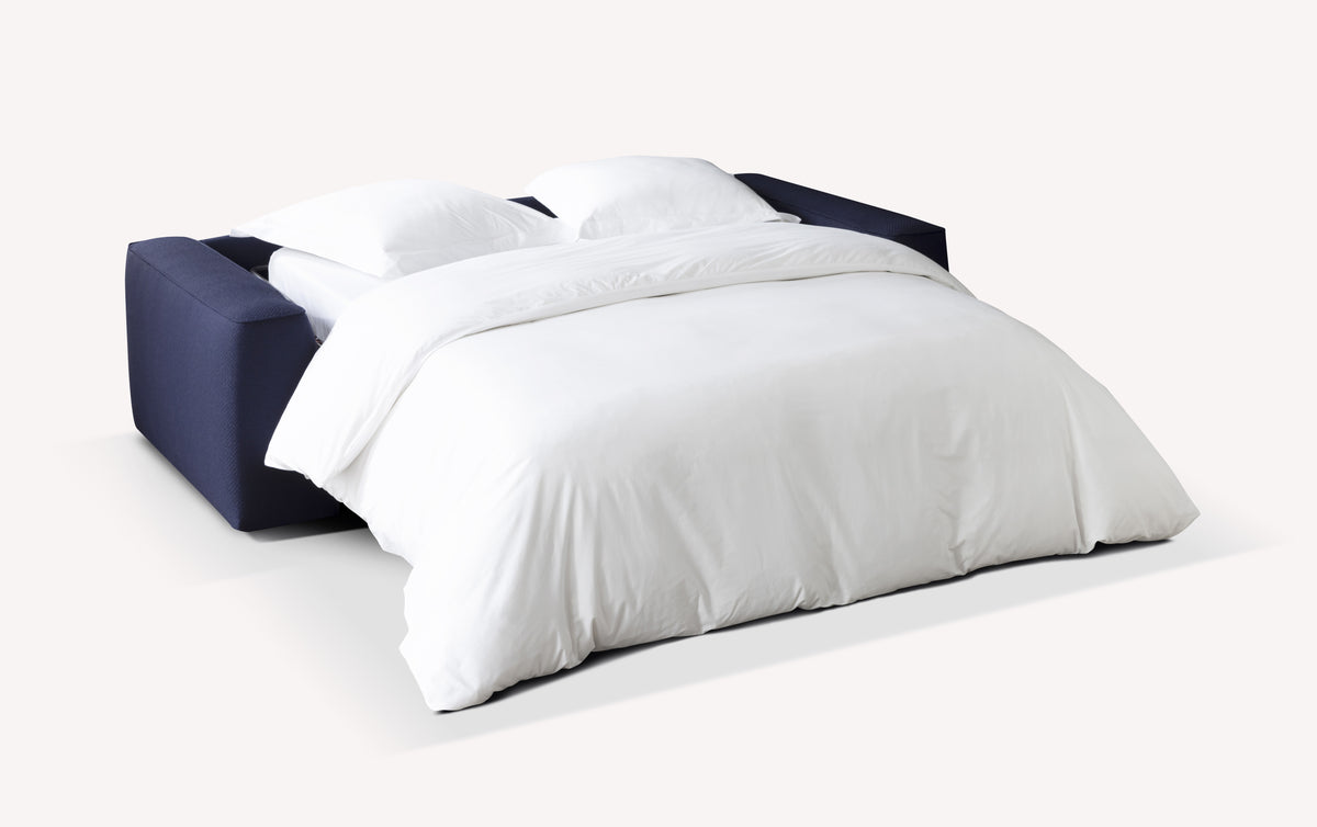 Lot canapé et pouf ONYX déplié en canapé convertible avec un couchage en tissu bleu foncé et une literie blanche, offrant un espace de couchage confortable pour un usage quotidien.