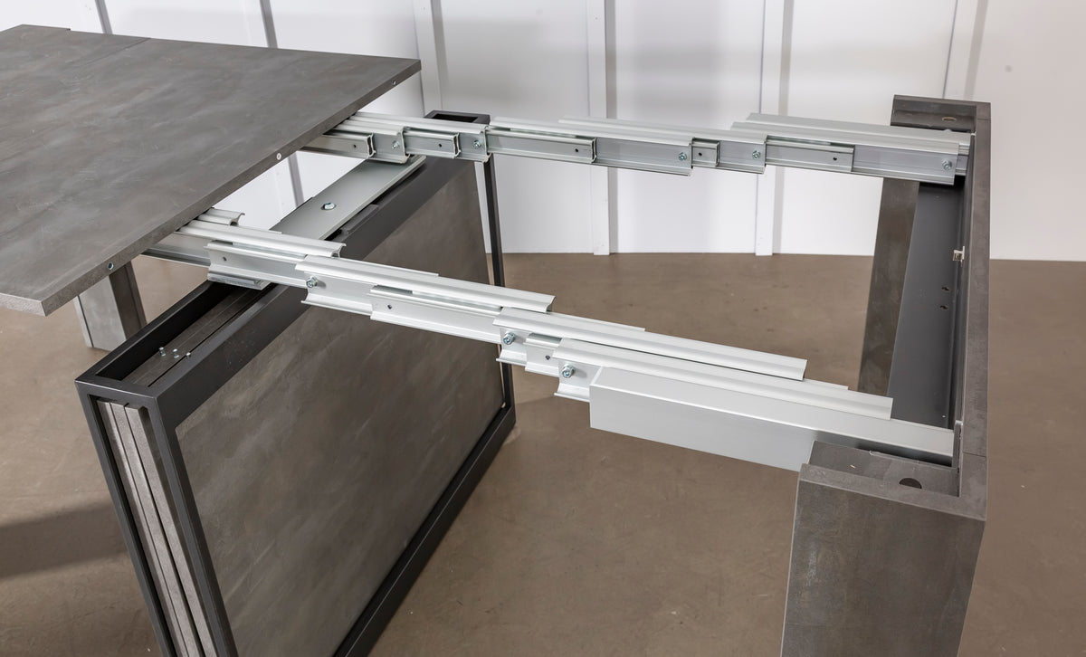 Table extensible ouverte montrant le mécanisme en métal sur un sol en béton, avec un plateau en COMPACT Mélaminé Pietra imitation pierre.