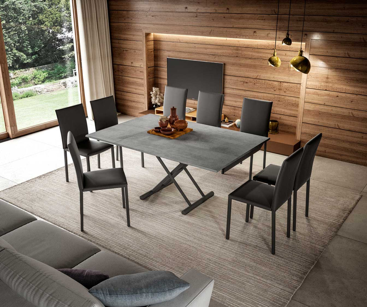 Salle à manger moderne avec table extensible PIU PIU Mélaminé Pietra en finition pierre, entourée de six chaises noires, ambiance chaleureuse avec mur en bois et éclairage doré.