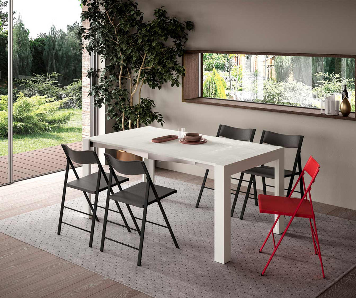 Table extensible SLIMMY Mélaminé Bianco spatolato dans une salle à manger lumineuse avec chaises pliantes noires et une rouge, décor minimaliste avec vue sur jardin verdoyant.