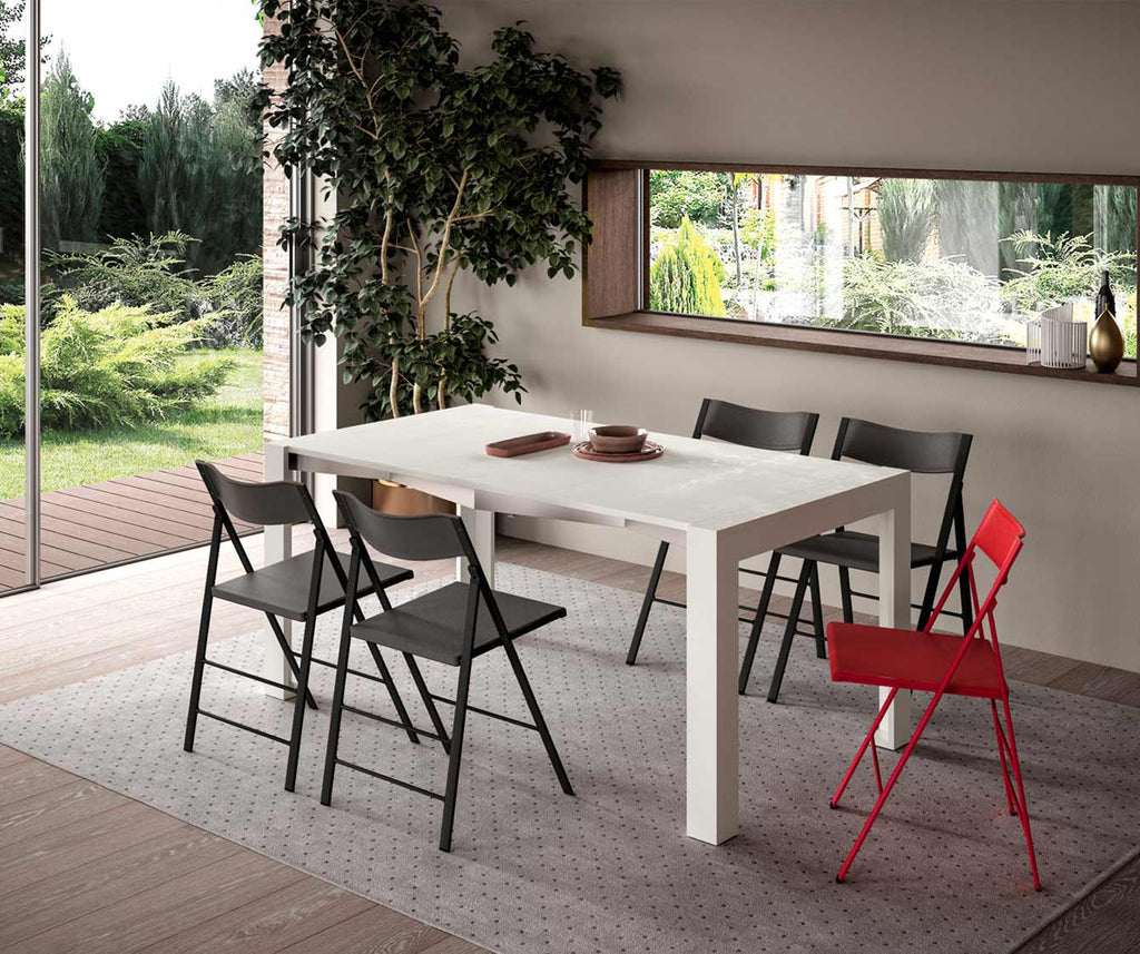 Table extensible SLIMMY Mélaminé Bianco spatolato dans une salle à manger lumineuse avec chaises pliantes noires et une rouge, décor minimaliste avec vue sur jardin verdoyant.