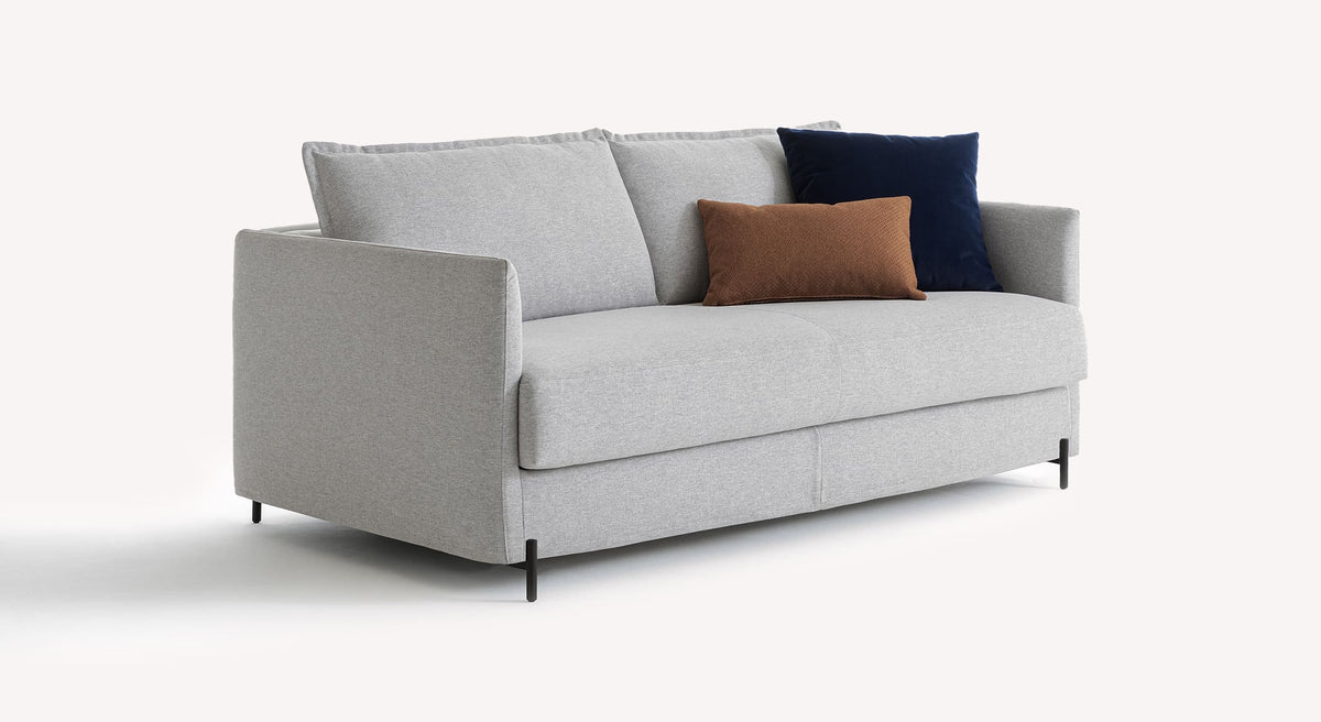 Canapé convertible SERGE Tixer Silver en tissu gris clair avec pieds métalliques noirs, décoré de coussins bleu marine et marron, idéal pour un couchage quotidien.