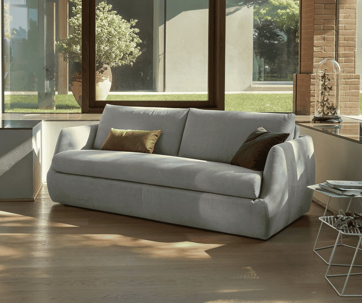 Canapé convertible LUZ Foster Silver en tissu gris clair, décoré de coussins, installé dans un salon lumineux avec parquet en bois et grandes fenêtres offrant une vue sur un jardin verdoyant.