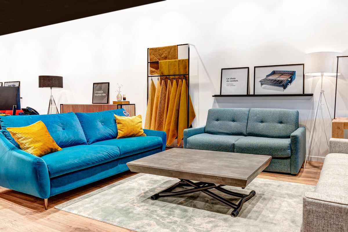 Salon moderne avec canapés bleus et coussins jaunes, table basse grise, et décorations murales, dans un showroom de La Maison du Convertible