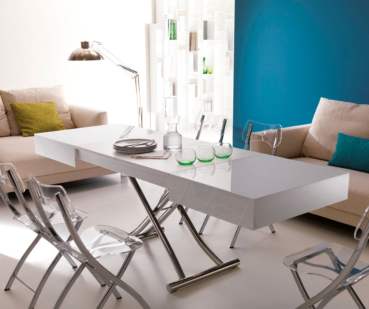 Table extensible moderne avec surface brillante blanc laqué accompagnée de chaises transparentes, mise en scène avec un décor chaleureux, illustrant le modèle BOX Verre Extrachiaro.