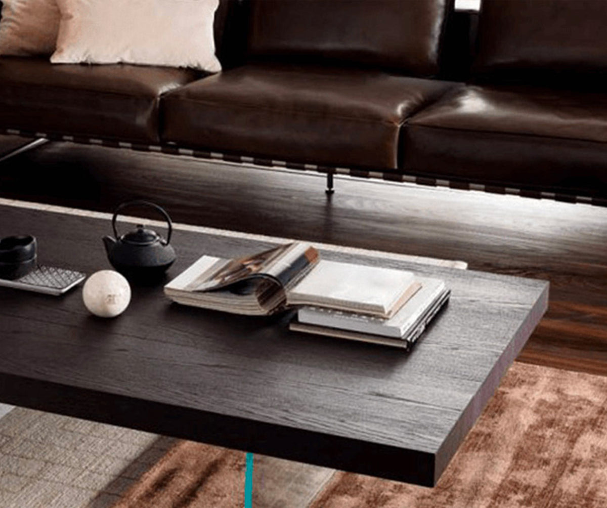 Table basse moderne en MARKUS Plaqué Rovere vecchio naturale, avec finition bois foncé, posée sur un tapis beige et marron, décorée de livres et d'une théière noire, devant un canapé en cuir.