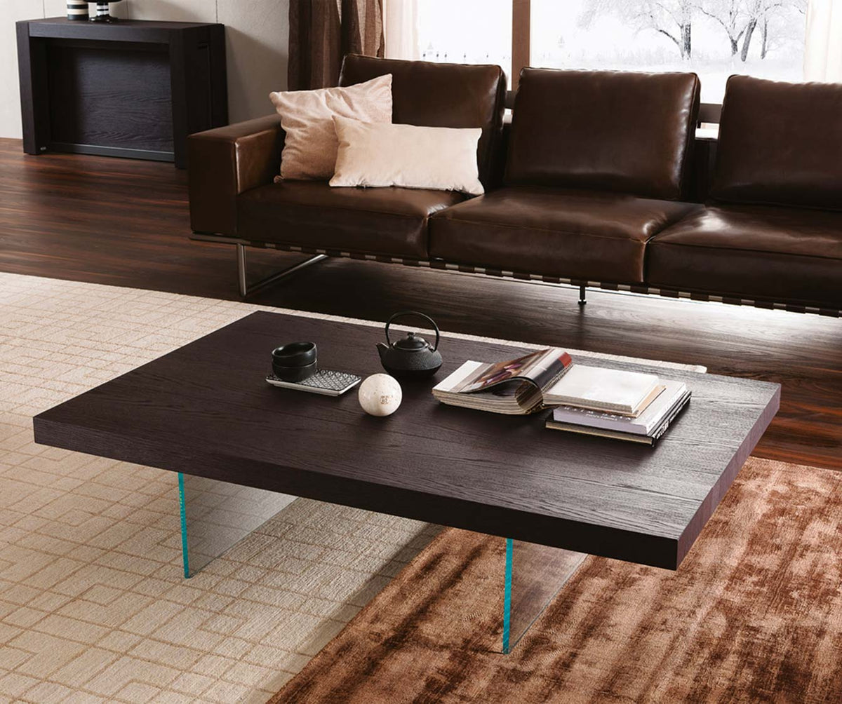 Table basse contemporaine MARKUS Plaqué Rovere vecchio naturale avec plateau en bois foncé et pieds transparents, posée dans un salon élégant avec canapé en cuir marron.