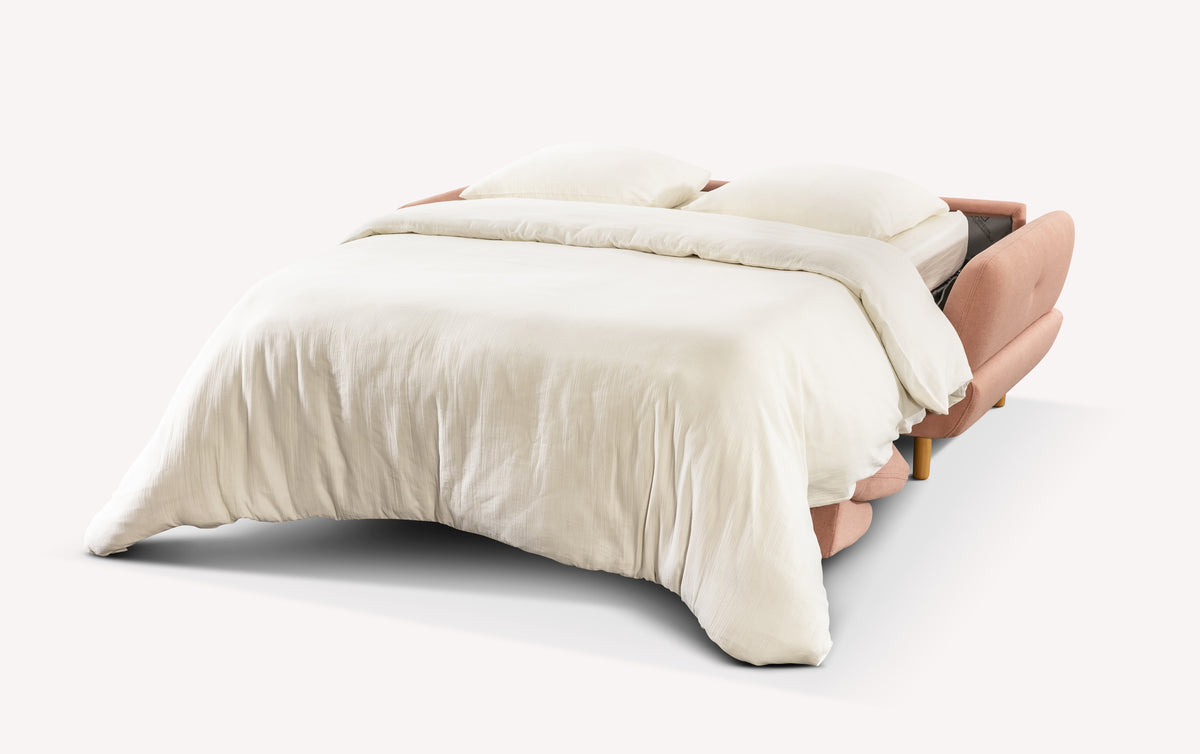 Canapé convertible ouvert en tissu rose, modèle Finn, avec draps blancs et coussins, sur fond blanc