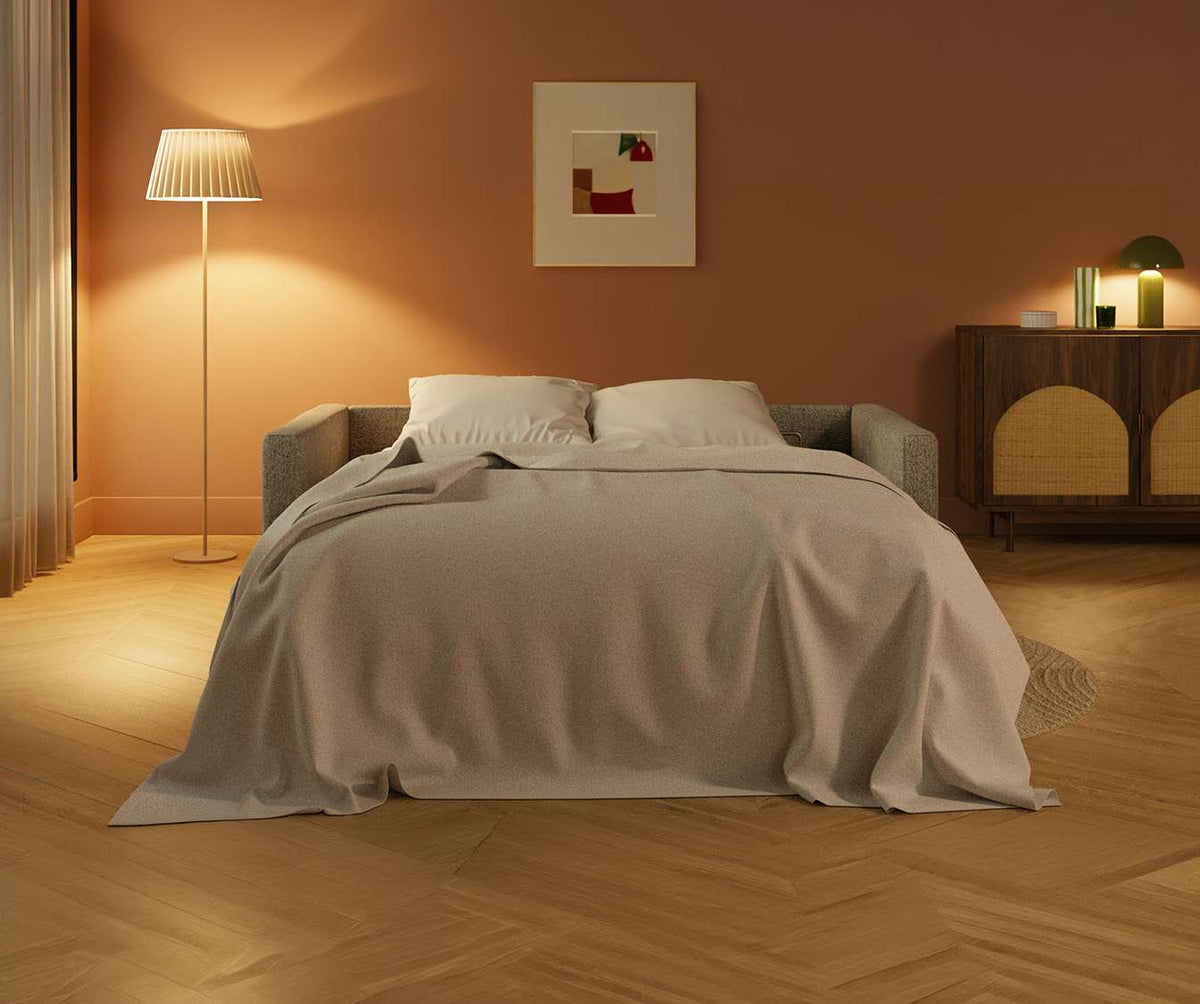 Canapé convertible JIMMY Foster Grey déployé en lit dans un salon chaleureux, habillé d’un tissu gris avec un plaid beige et deux coussins blancs, ambiance cosy et élégante.