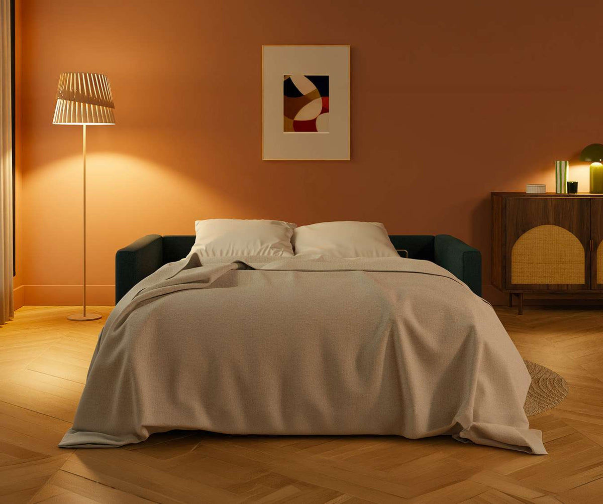 Canapé convertible JIMMY Foster Bleu ocean déplié en couchage avec couverture beige dans un salon chaleureux au mur terracotta, éclairé par une lampe sur pied.