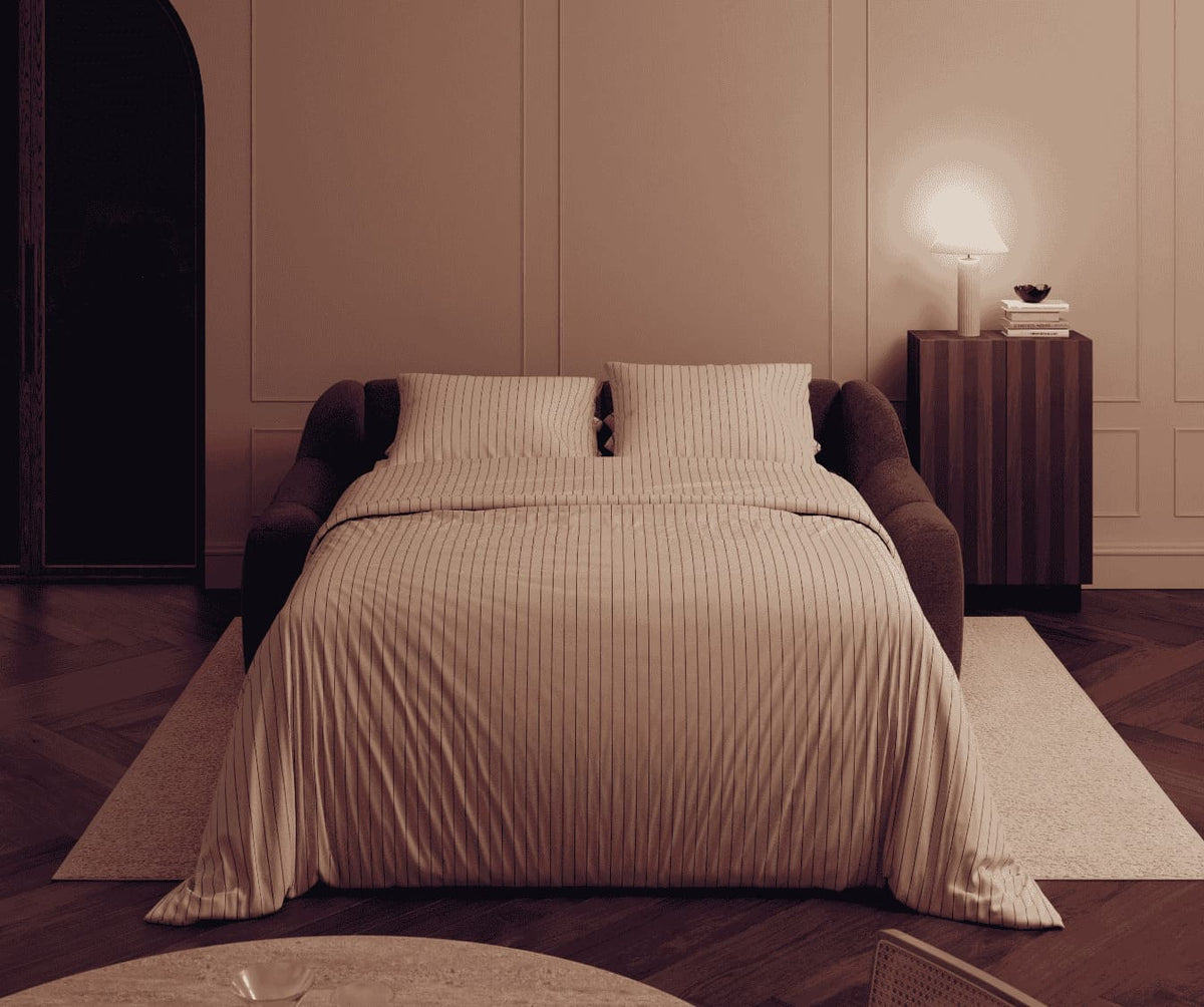 Canapé convertible MAMA Sogno Ocean déplié en lit, dans une chambre élégante avec linge de lit rayé beige, ambiance chaleureuse et mobilier design.