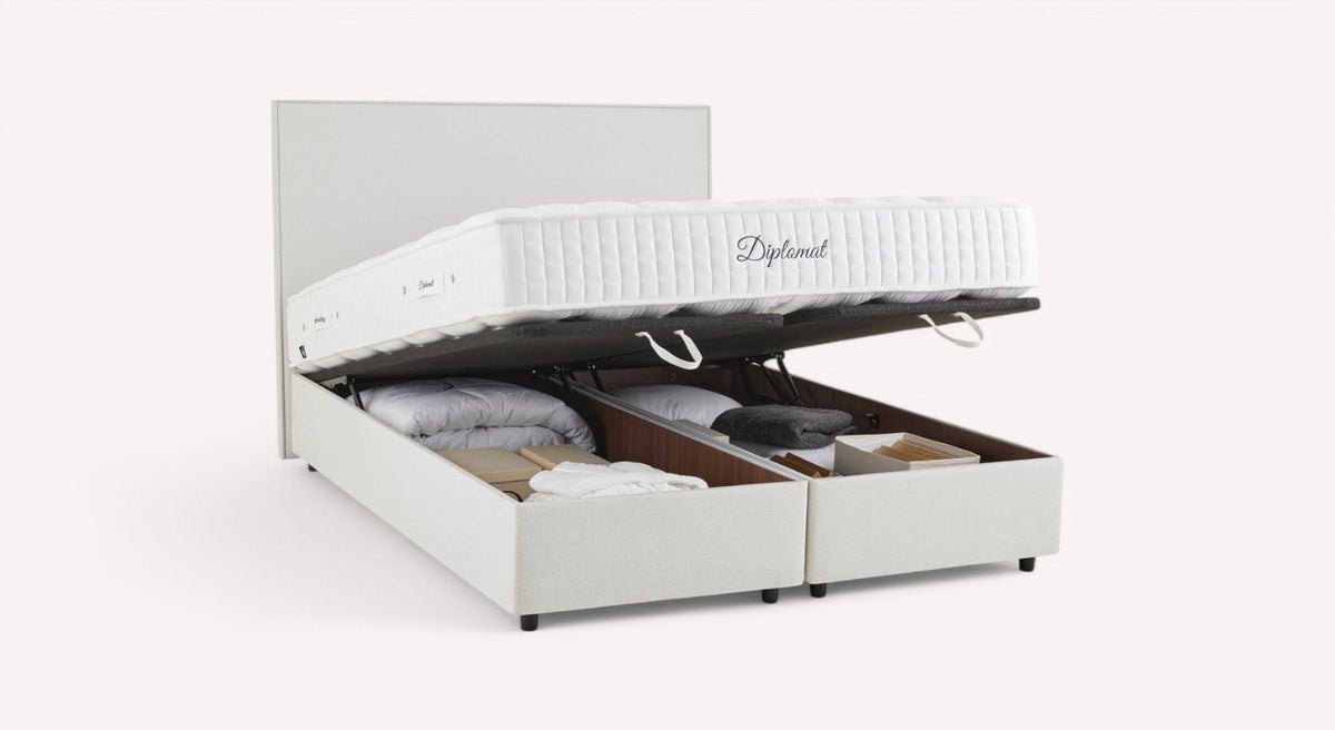 Lit coffre blanc avec matelas "Diplomat" relevé, montrant un espace de rangement contenant des couvertures et des boîtes