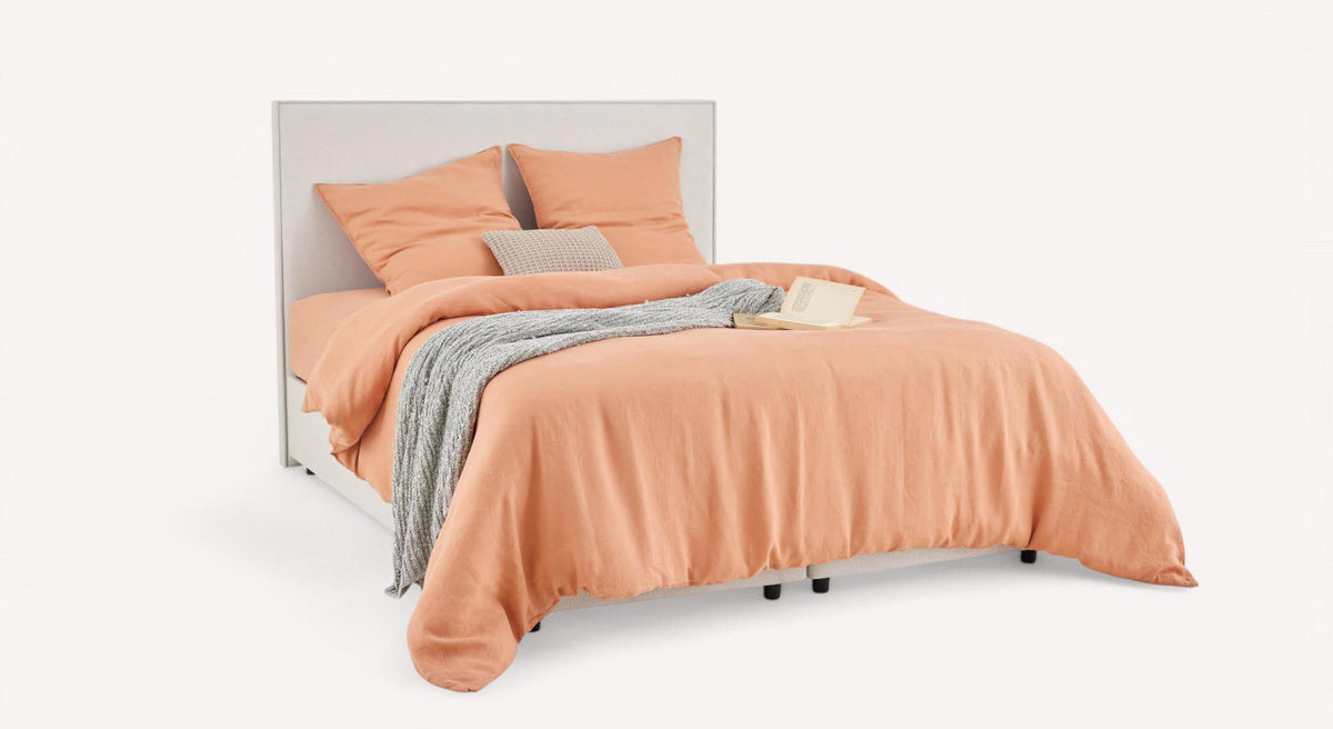 Lit avec housse de couette pêche, coussins assortis, couverture grise et livre ouvert, sur fond blanc