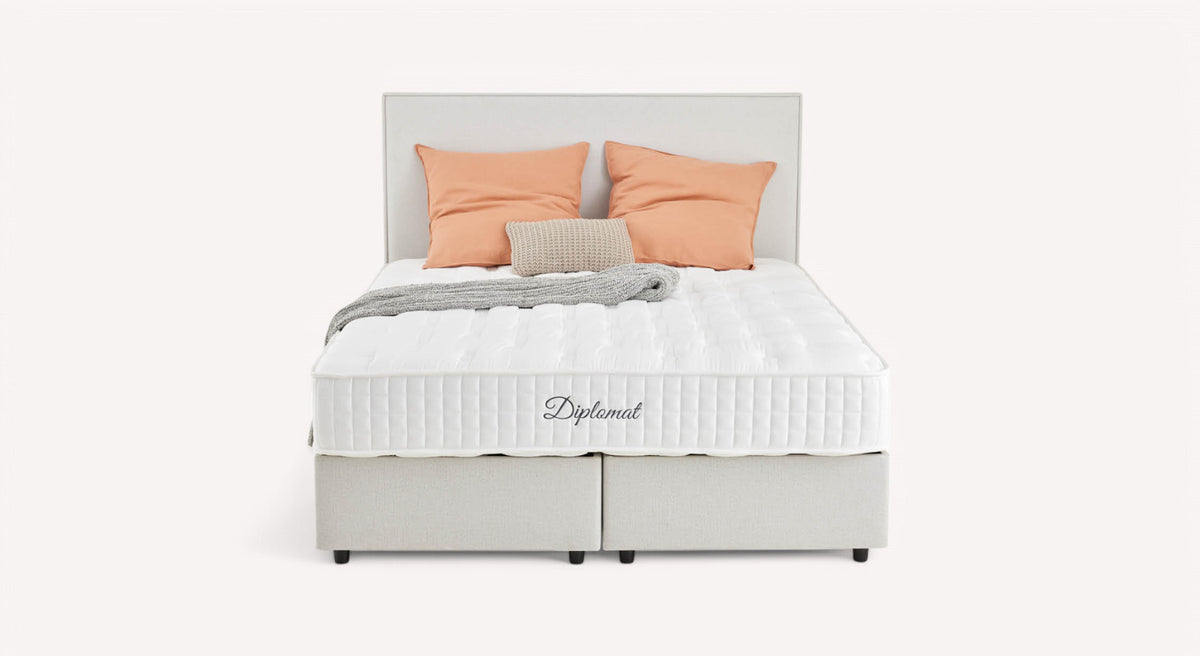 Lit avec matelas blanc "Diplomat", deux oreillers orange, un coussin beige et une couverture grise, sur fond blanc