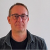 Homme portant des lunettes et une chemise grise, debout devant un fond uni